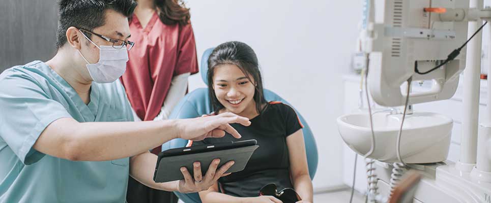 Doctor in mask showing a tablet to a smiling patient in a dental office.