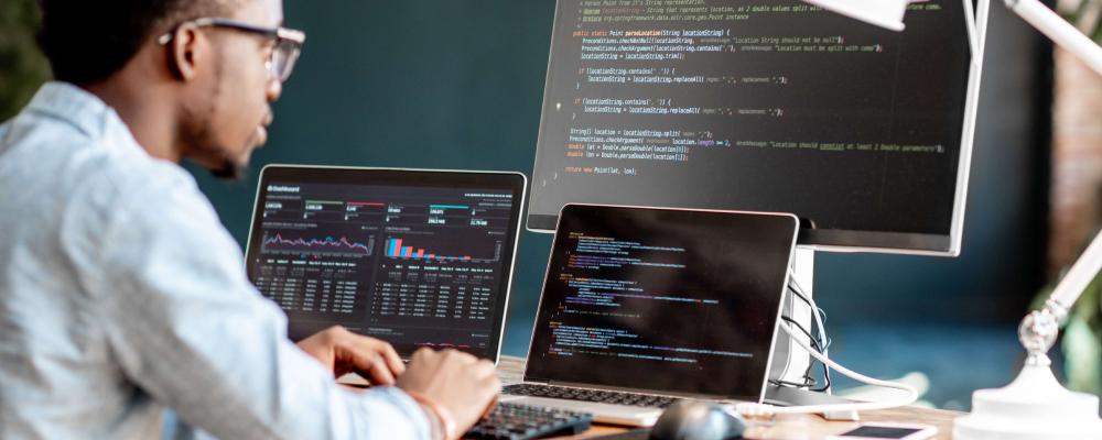 Black man in glasses working on a laptop, second laptop, and monitor displaying code/charts.