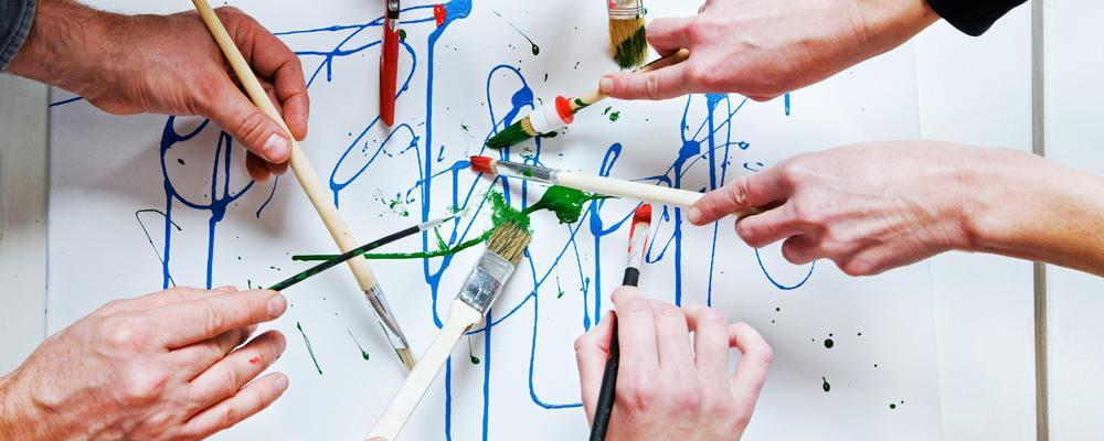 Multiple hands paint with brushes and colorful paints on a white surface.