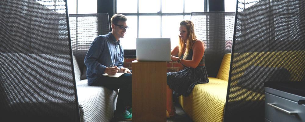 Two people on yellow benches, working on a laptop at a small table near large windows.