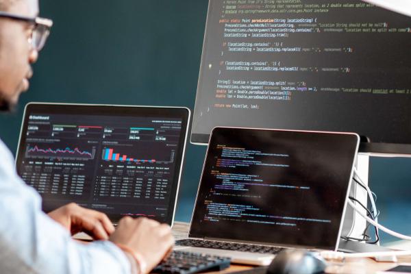 Black man in glasses working on a laptop, second laptop, and monitor displaying code/charts.