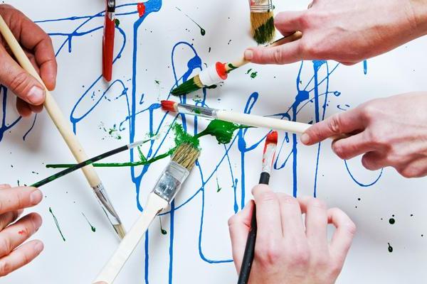 Multiple hands paint with brushes and colorful paints on a white surface.