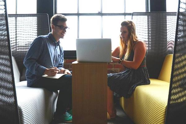 Two people on yellow benches, working on a laptop at a small table near large windows.